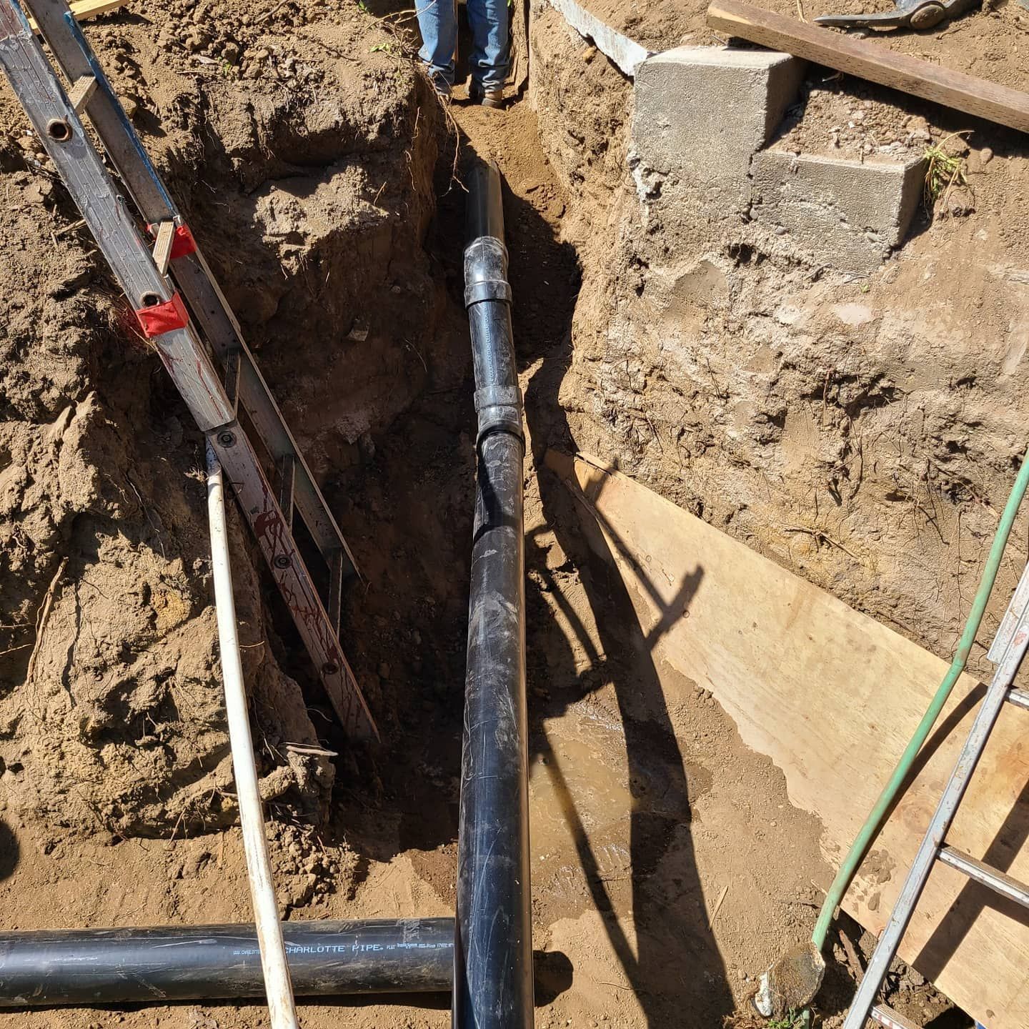 Black pipes in a dirt trench, construction site; ladder, wood planks, and person visible.