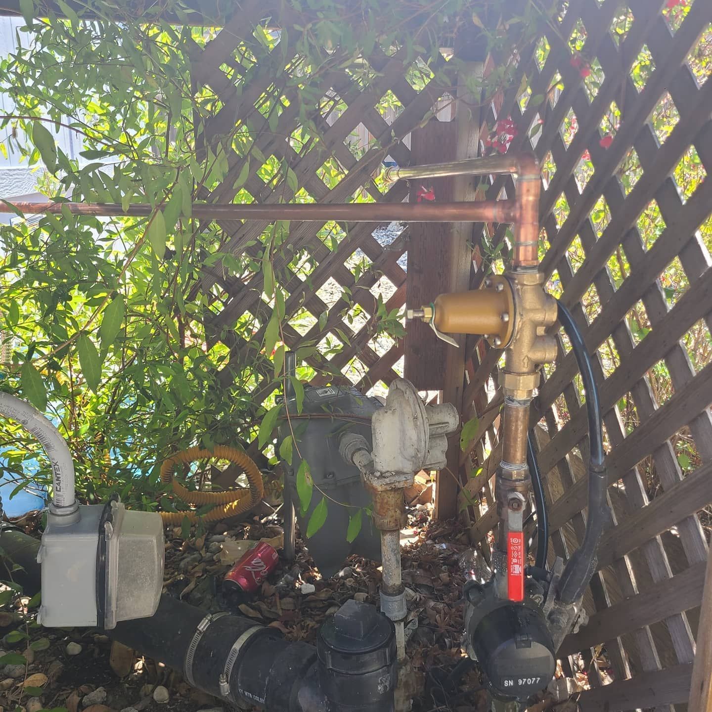 Copper pipes, valve, and gas meter behind a wooden trellis, with foliage.