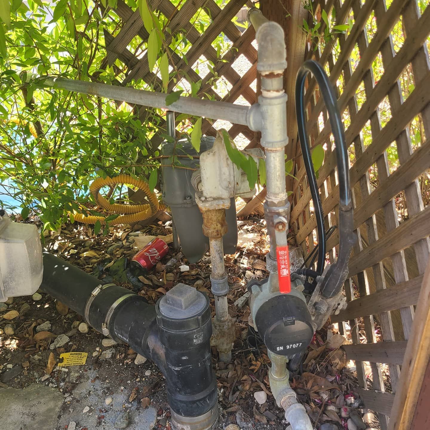 Water meter and pipes behind a lattice fence, with some greenery visible.