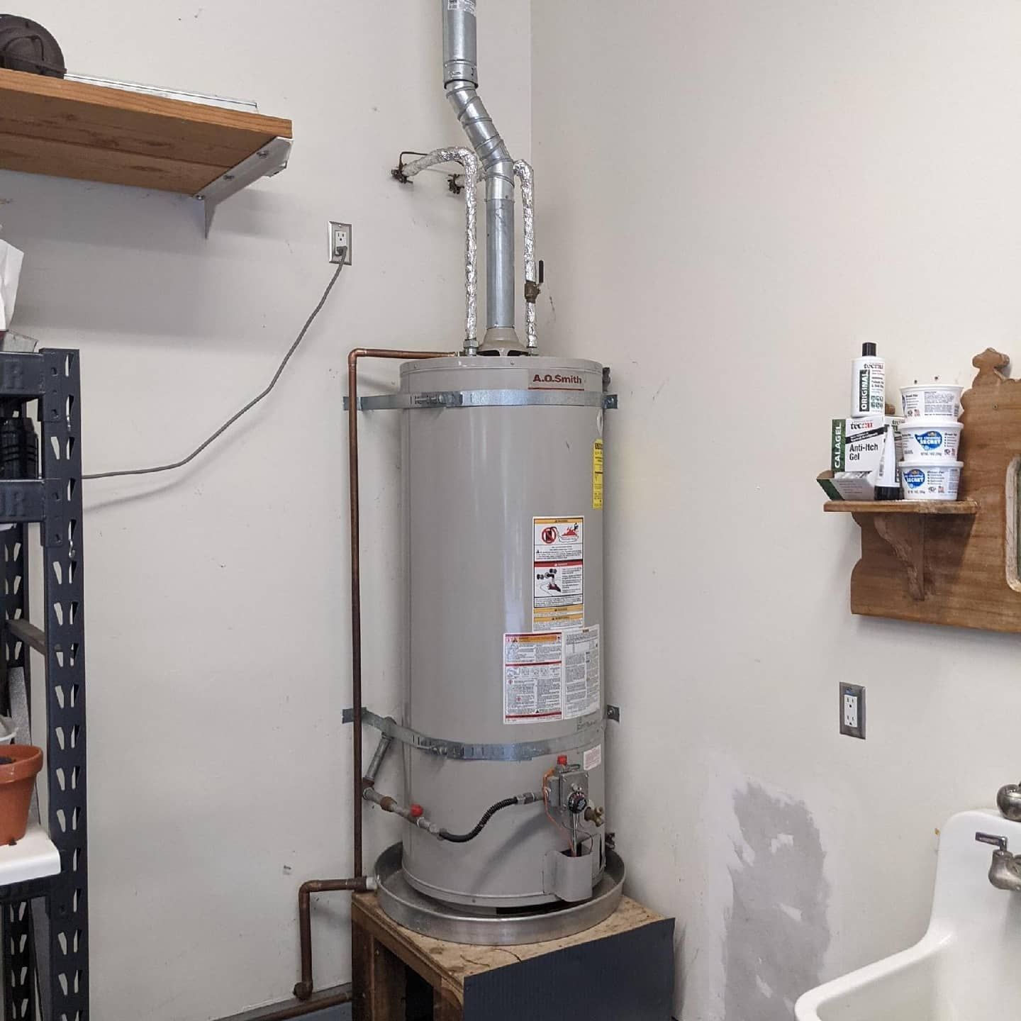 A gas water heater, silver and tall, stands against a white wall in a utility room. Copper pipes and vent are visible.
