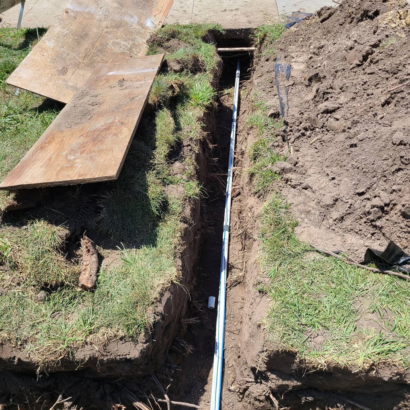Trench dug in grass, with electrical conduit and wires running through it. Shovel and wood plank nearby.