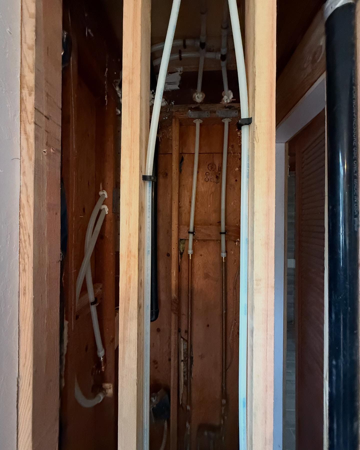 Open wall cavity with white PEX water pipes and black vent pipe. Wood studs and framing visible.