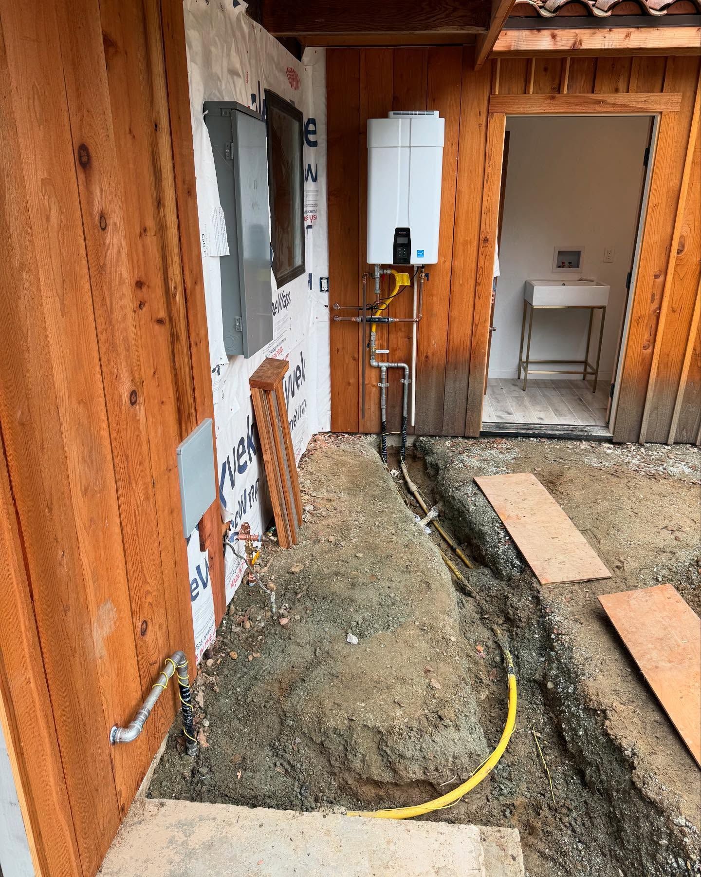 Exterior view: gas water heater mounted on a wooden wall, trench with yellow pipe, wooden boards, and a door to a room.