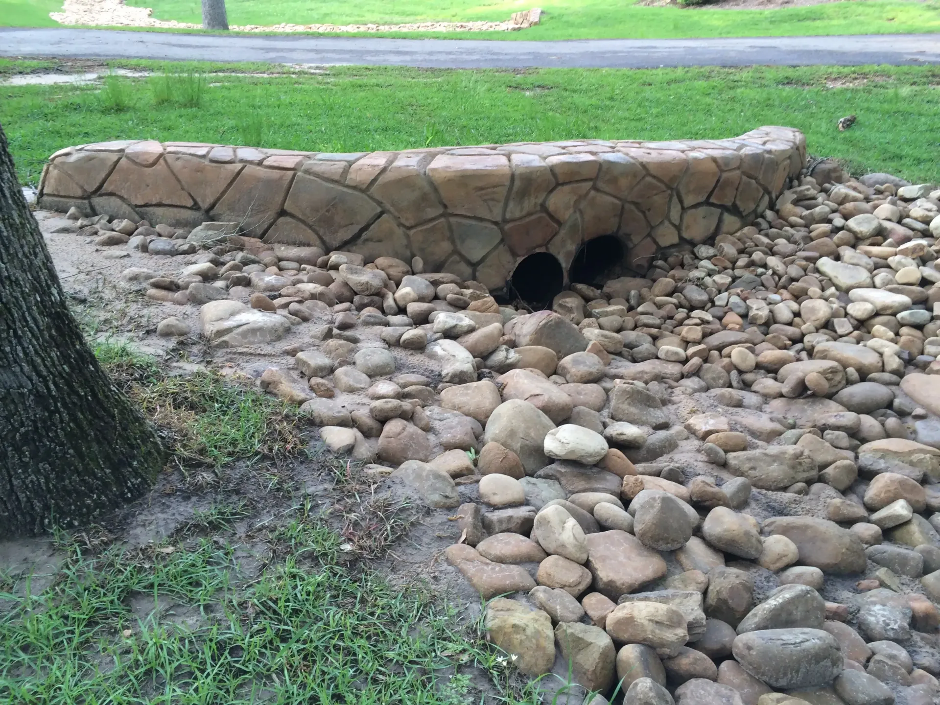Stone culvert with two dark openings, surrounded by rocks and a stone wall, in a grassy area.