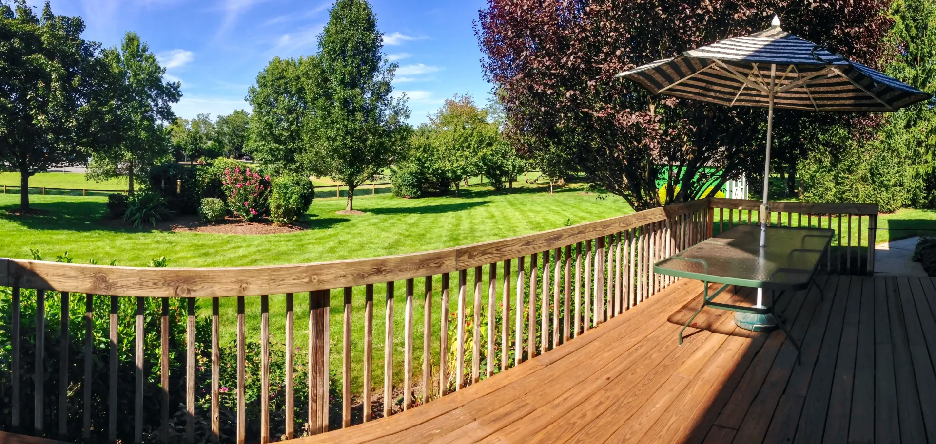 Wooden deck overlooking a green lawn with trees and an umbrella-covered table. Sunny, blue sky.