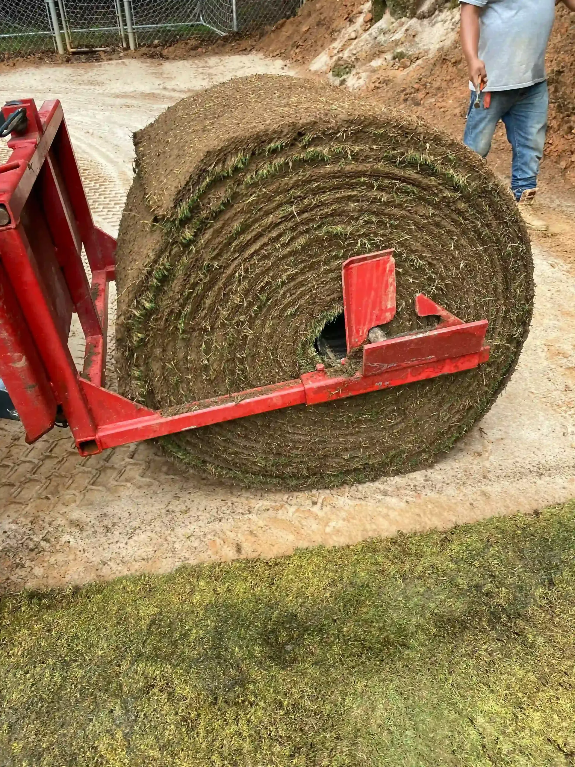 Large roll of sod being transported by a red machine on a light-colored surface with a person in the background.