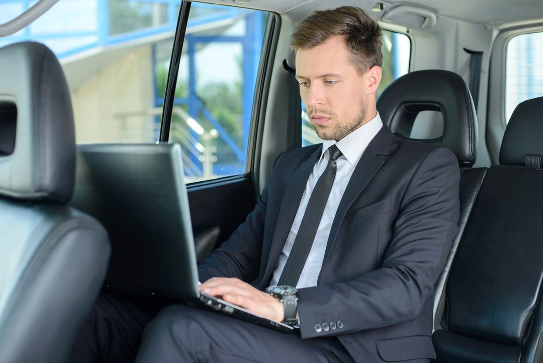 A man in a suit is sitting in the back seat of a car using a laptop computer.