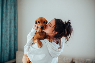 Woman in white hoodie kisses a brown dog, indoors.