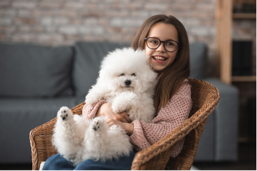 Girl with glasses smiles, holding a fluffy white dog in her lap, seated in a wicker chair.