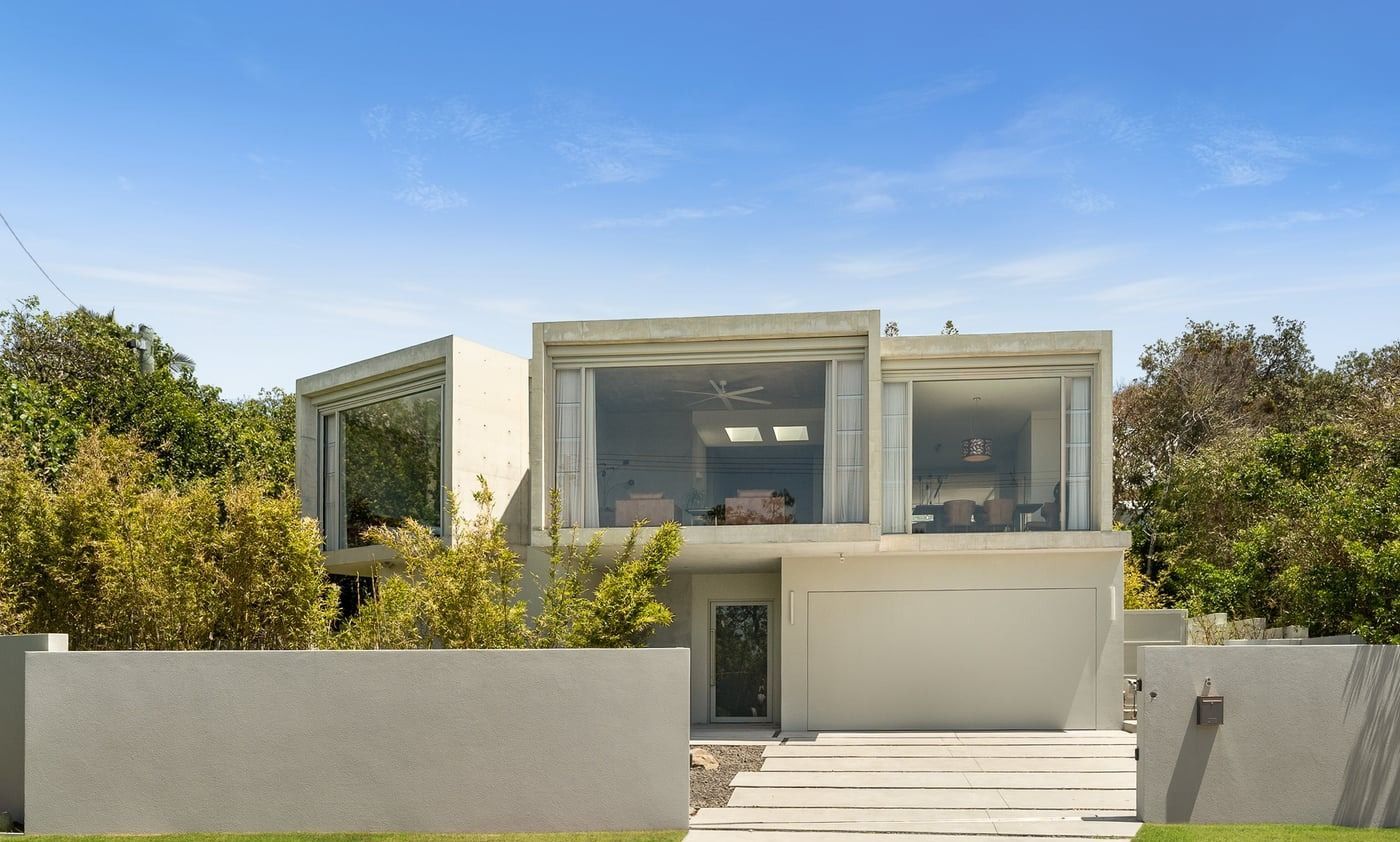 Modern, light-colored concrete two-story house with large glass windows, surrounded by trees and a perimeter wall — SC Glass & Aluminium in Yandina, QLD