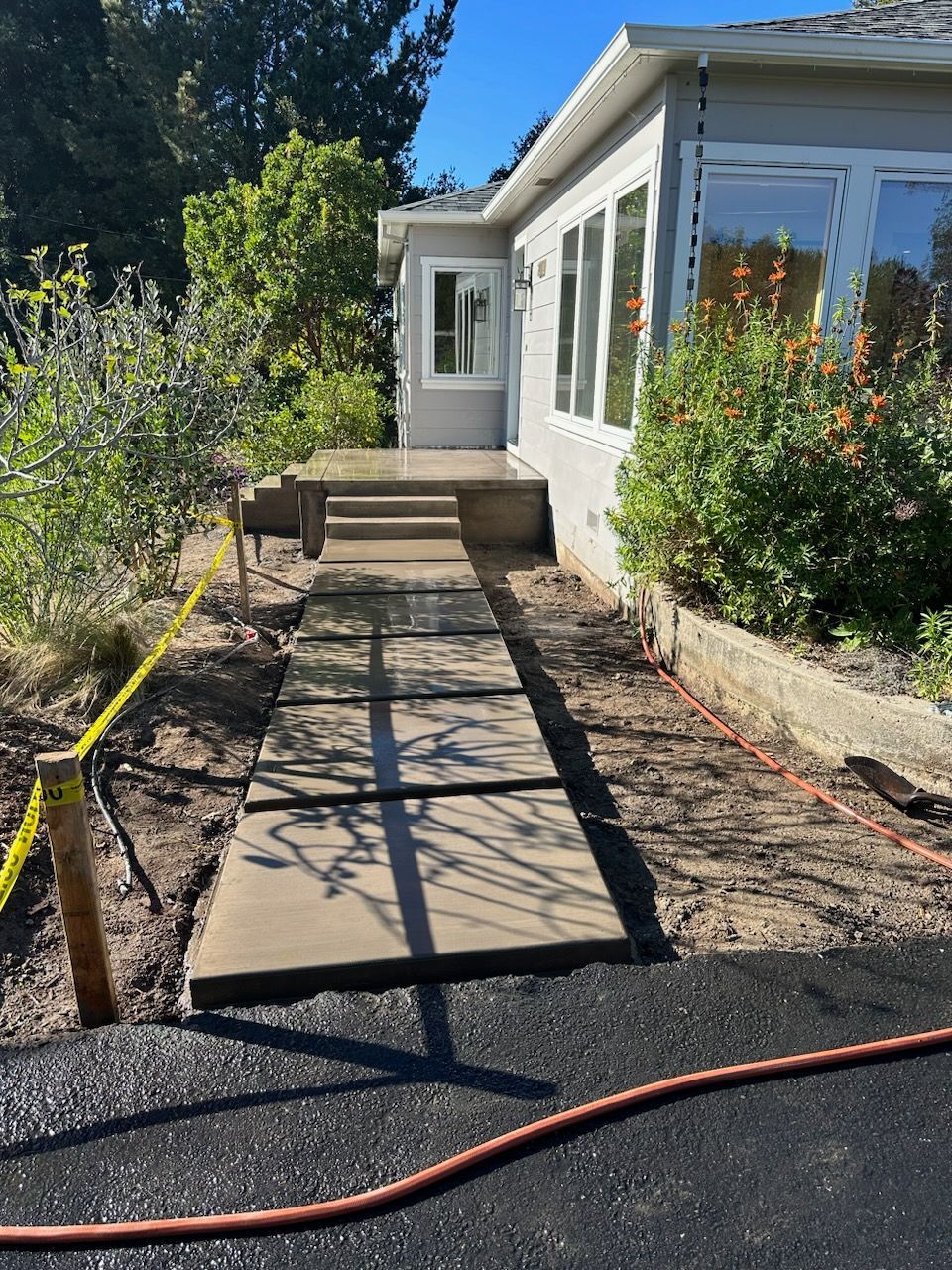 Concrete pathway leading to a house with large windows, set in a sunny, outdoor environment.