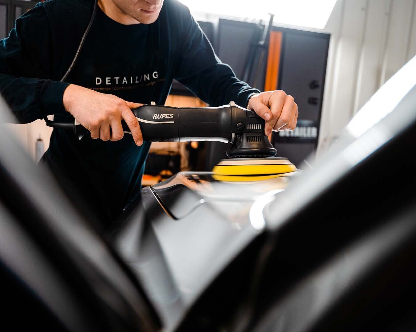 Person polishing a black car with an electric buffer, in a detailing studio.