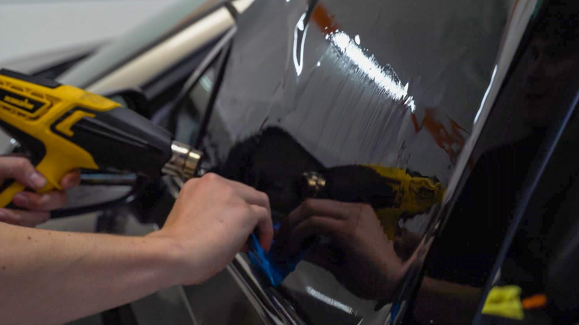 Person using a heat gun and smoothing tool to apply window tint to a black car.
