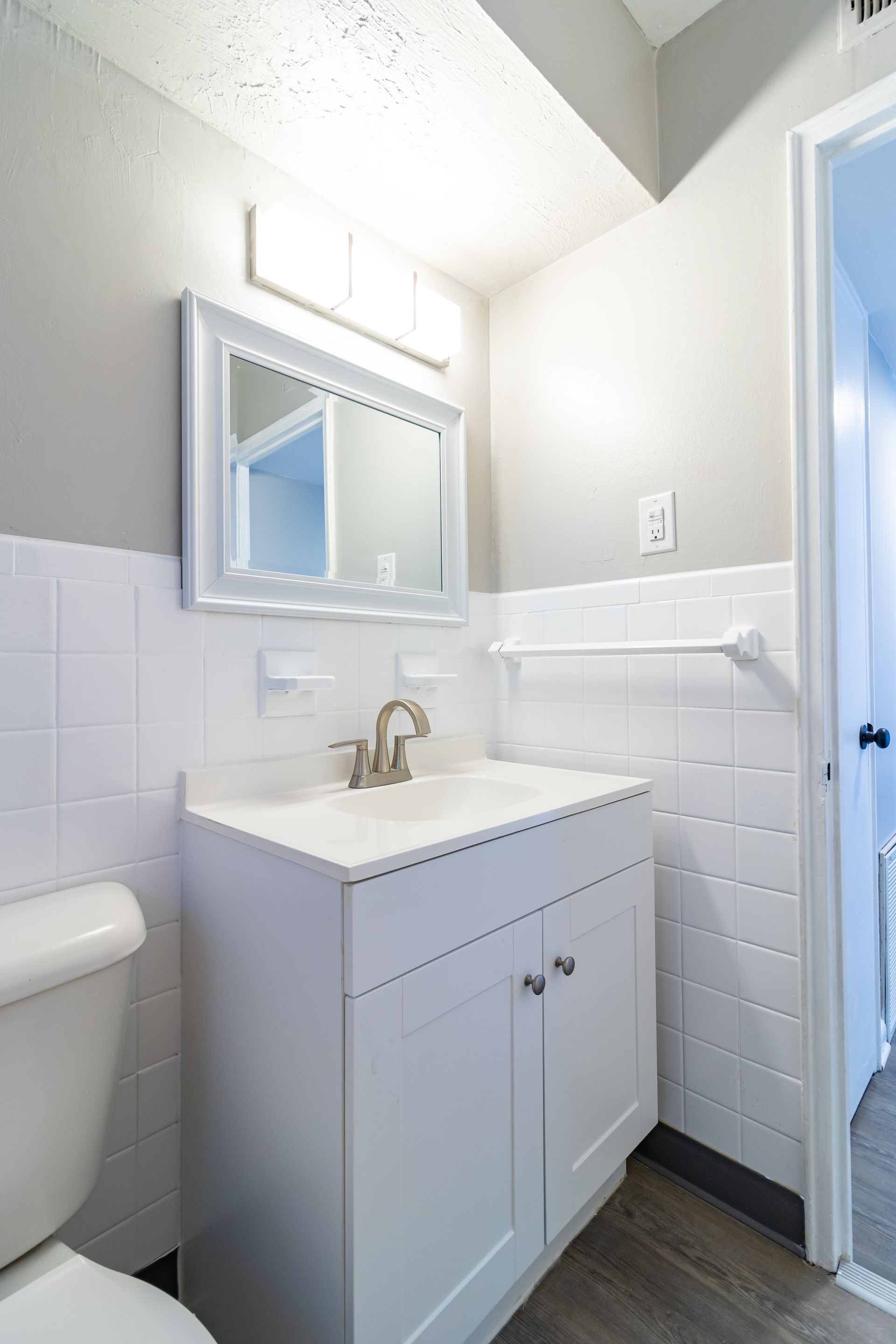 White bathroom with a sink, mirror, and toilet. Tile on the walls and a partially open doorway.