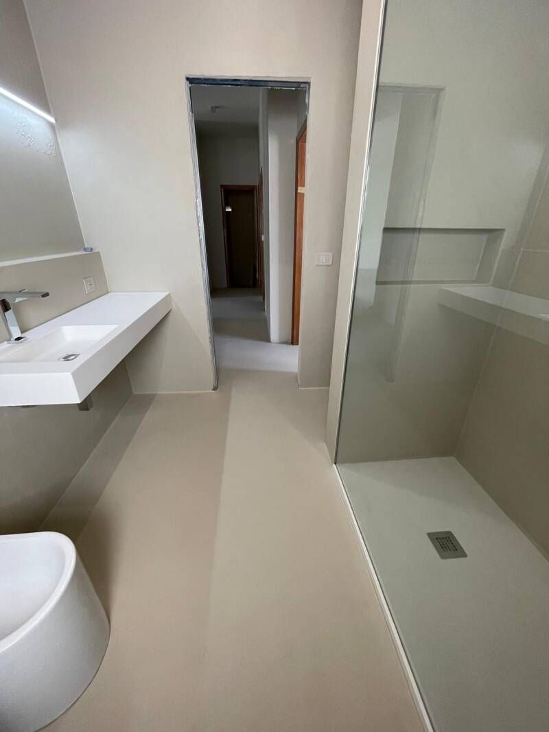 Bagno moderno con lavandino, doccia e WC bianchi. Pavimenti e pareti beige chiaro.