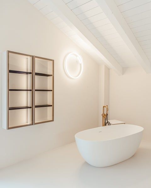 Moderno bagno bianco con vasca da bagno, mobiletto decorativo a muro e una luce circolare luminosa.
