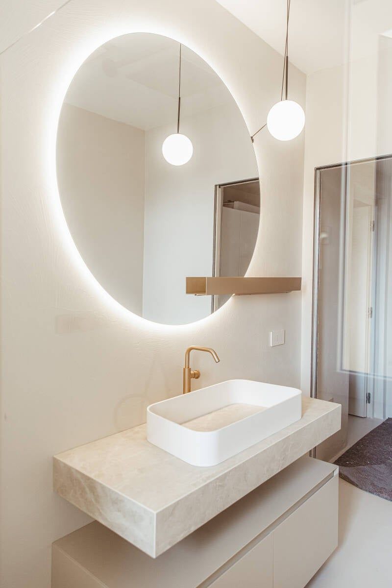 Bagno moderno con specchio ovale luminoso, lavandino rettangolare, rubinetto dorato e mobiletto sospeso.