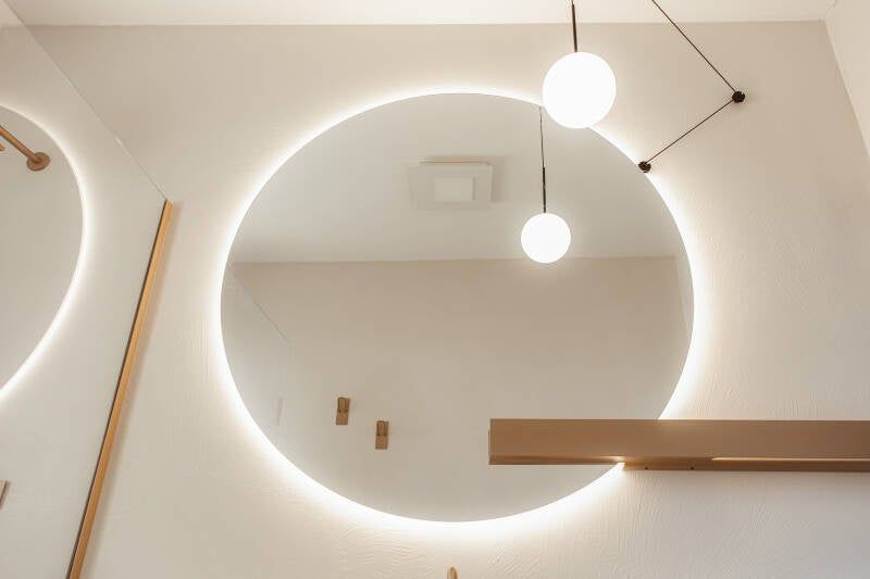 Specchio circolare con bordo illuminato, due lampade a globo sospese e mensola in legno su parete bianca.