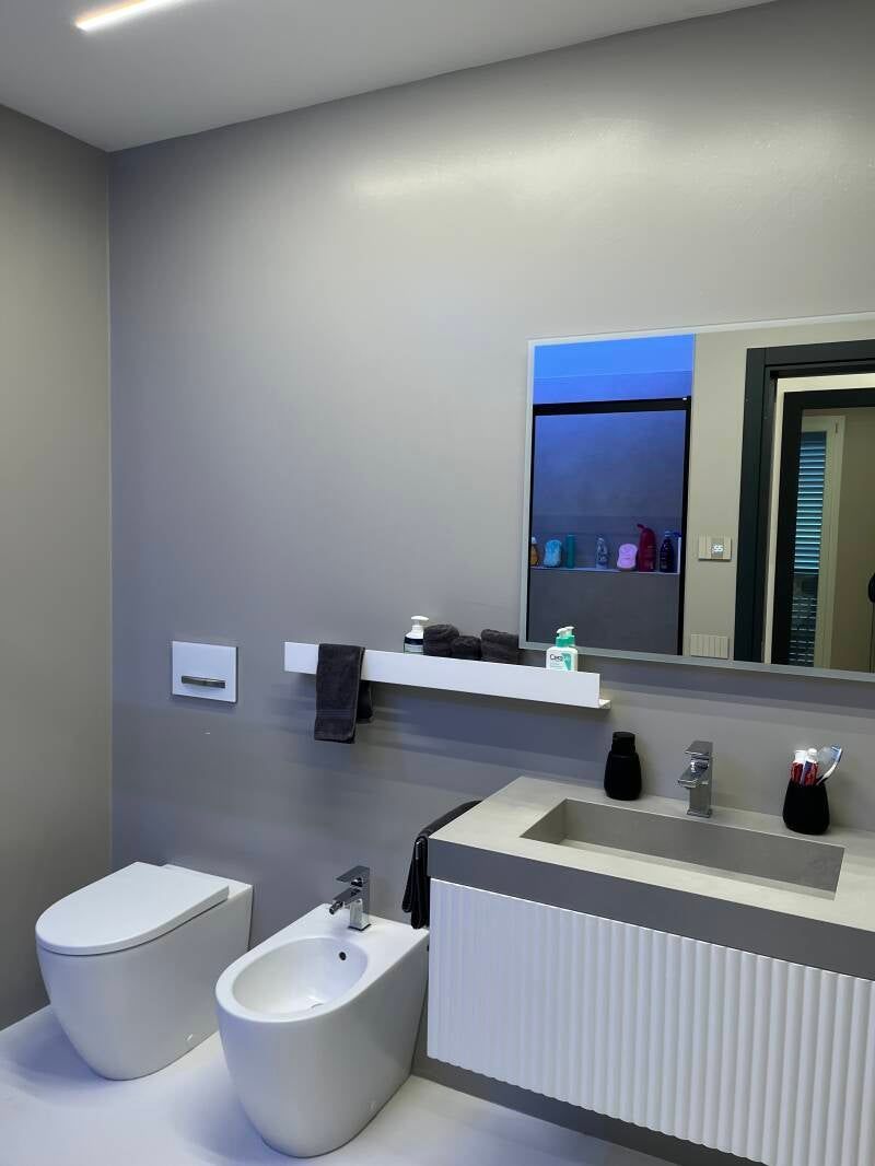 Bagno moderno con pareti grigie, WC, bidet, mobiletto con specchio e mensola bianca con oggetti.