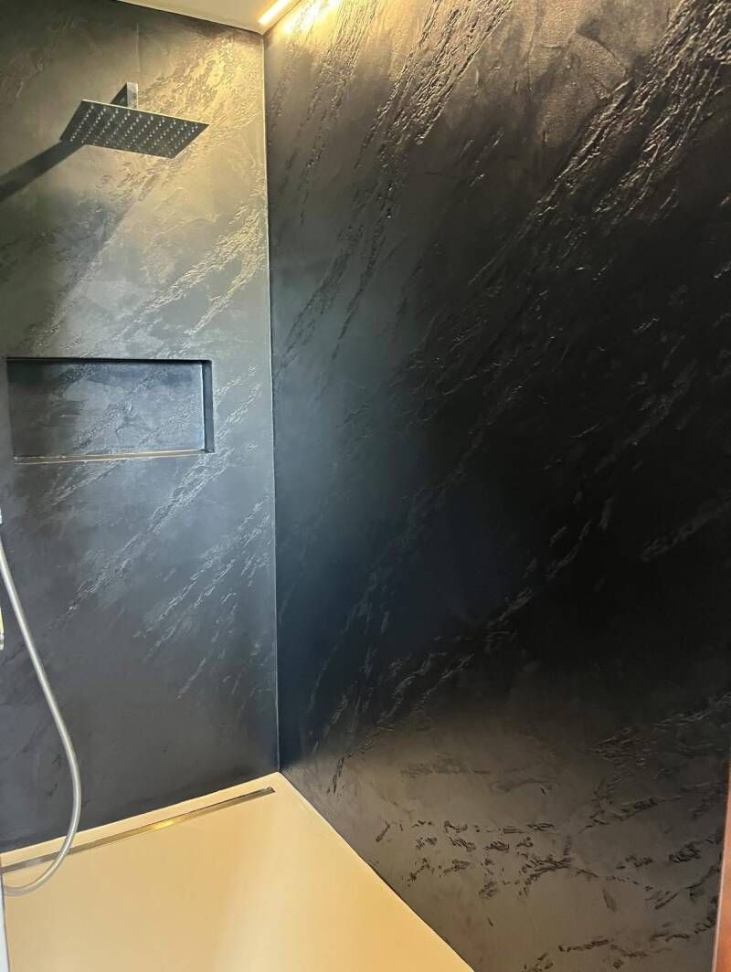 Doccia scura e testurizzata con mensola integrata e soffione quadrato, in un angolo. Pavimento beige.