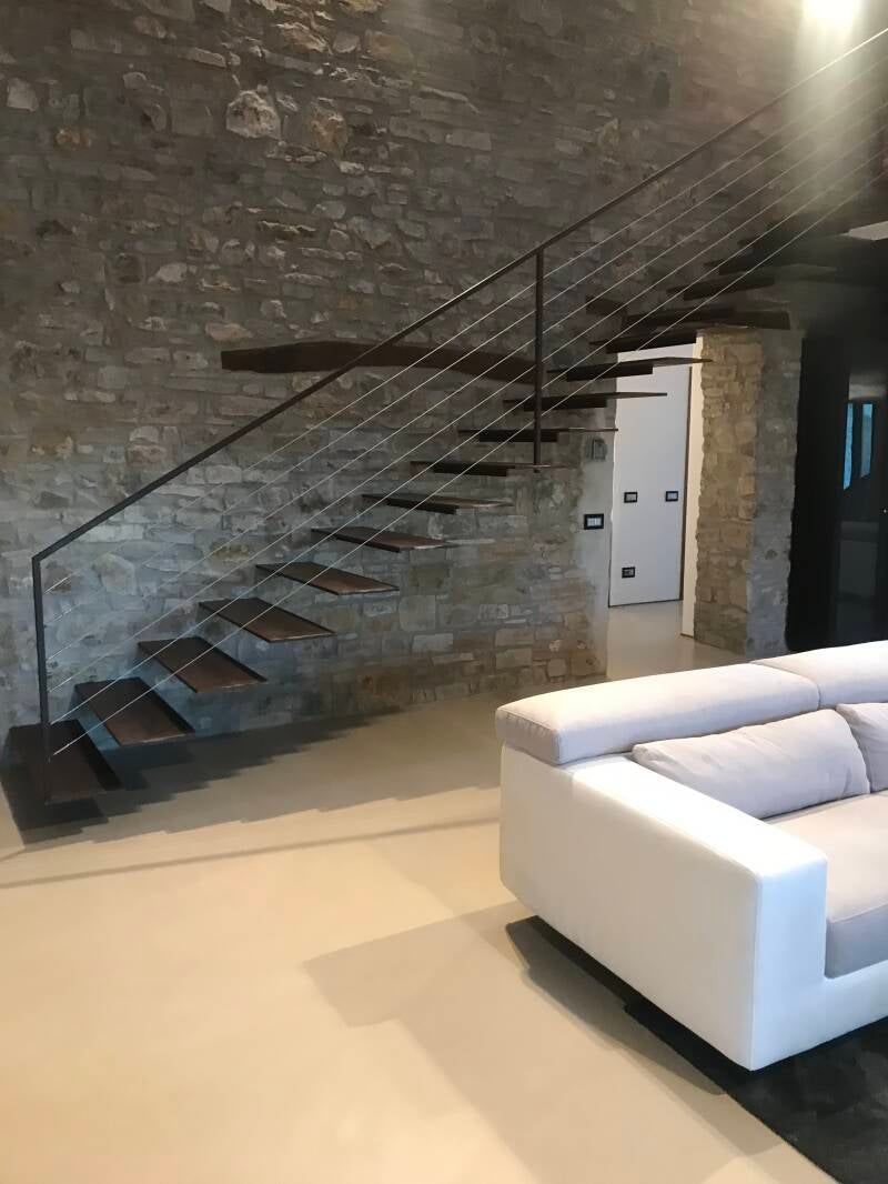 Scala sospesa in legno con corrimano in metallo contro un muro di pietra, accanto a un divano bianco.