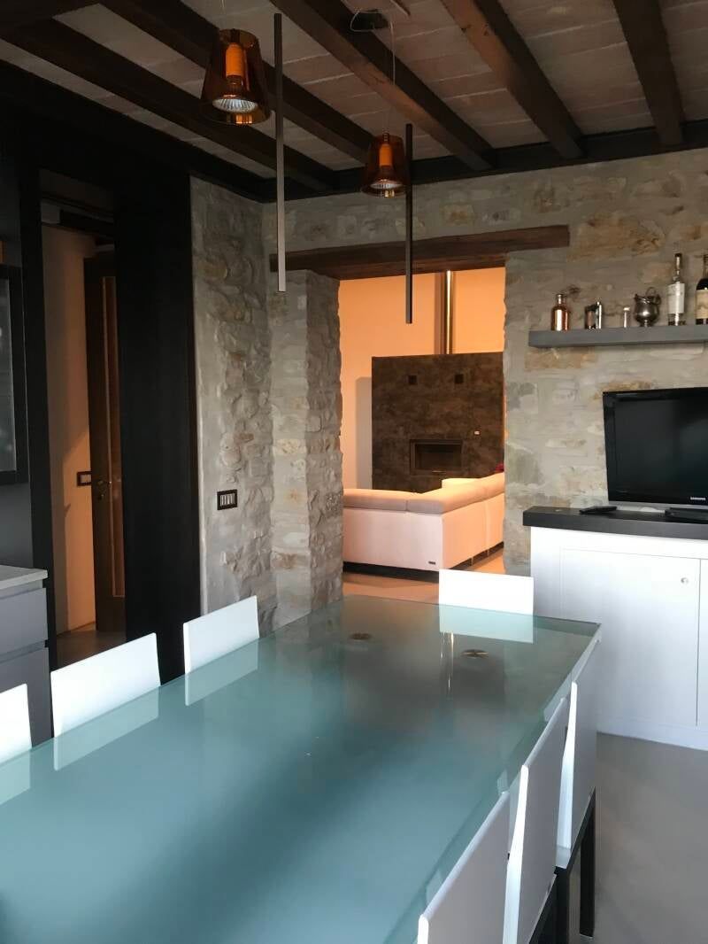 Sala da pranzo con tavolo in vetro smerigliato, pareti in pietra e vista sul soggiorno con divano bianco.