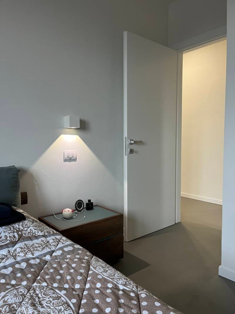 Interno della camera da letto con comodino, lampada, porta aperta su un'altra stanza.