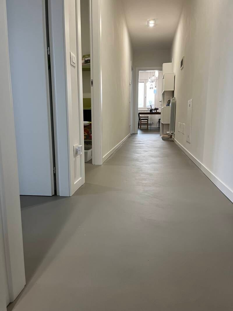 Corridoio stretto con pavimento grigio, pareti e porte bianche.