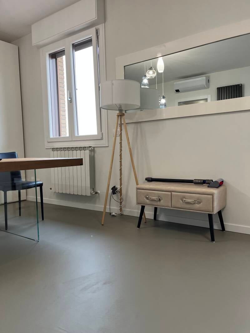 Stanza con una lampada da terra in legno, un piccolo mobiletto e un grande specchio che riflette la stanza.