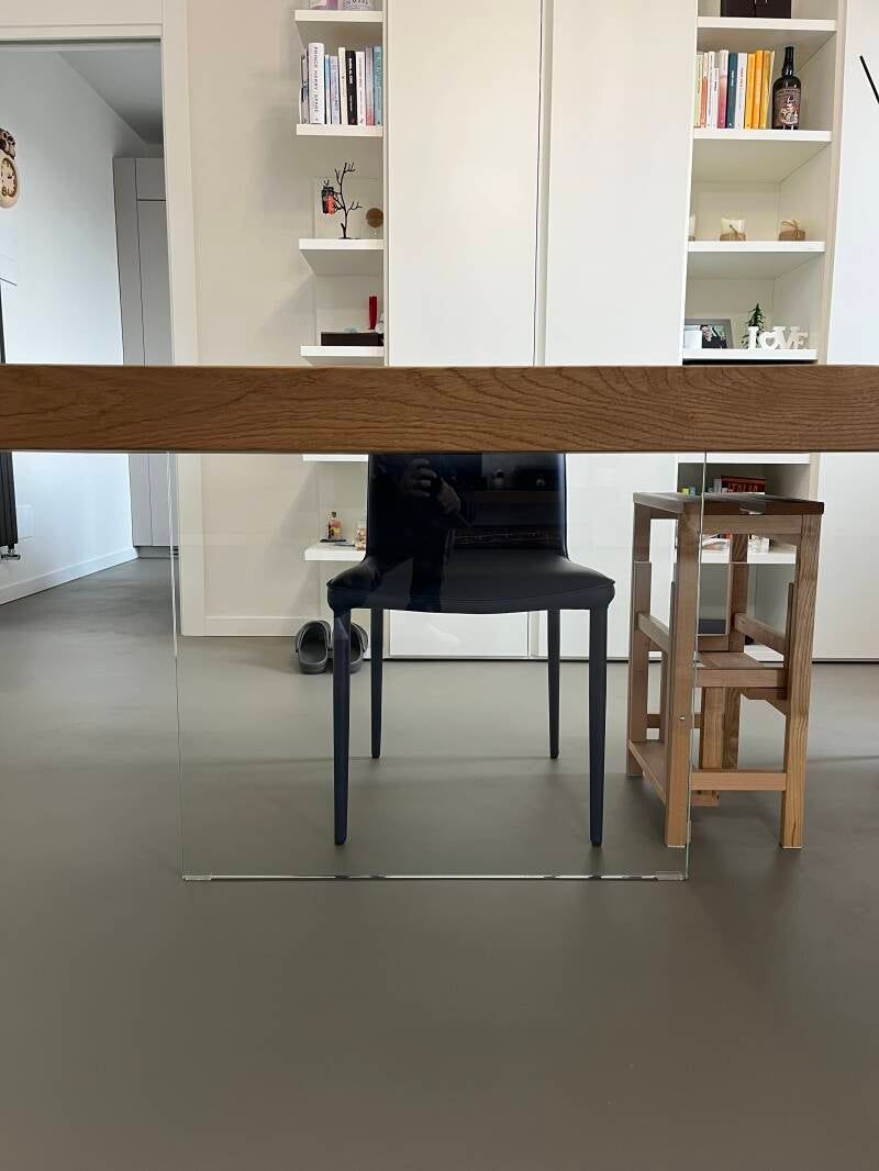 Una sedia scura e uno sgabello di legno sono posizionati sotto un tavolo di legno, sullo sfondo di una libreria bianca.