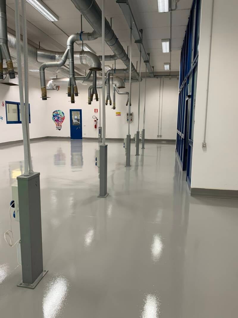 Interno di un laboratorio con pavimento grigio, tubi sospesi e numerosi pali di supporto argentati.