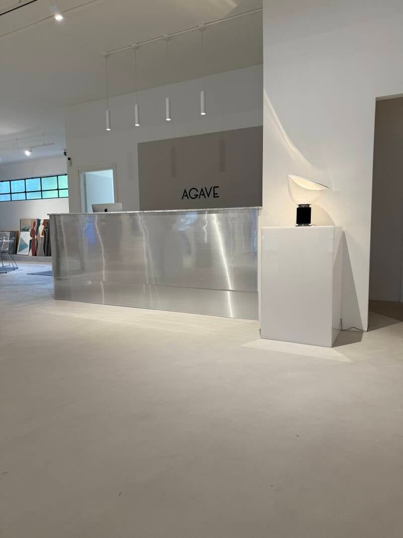 Area reception minimalista con scrivania argentata, pareti bianche e alcune luci sospese.