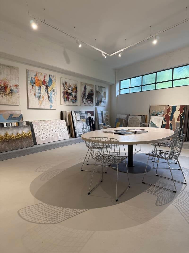 Interno di una galleria d'arte con dipinti esposti, tavolo ovale e sedie moderne. Spazio luminoso e arioso con dettagli dipinti sul pavimento.