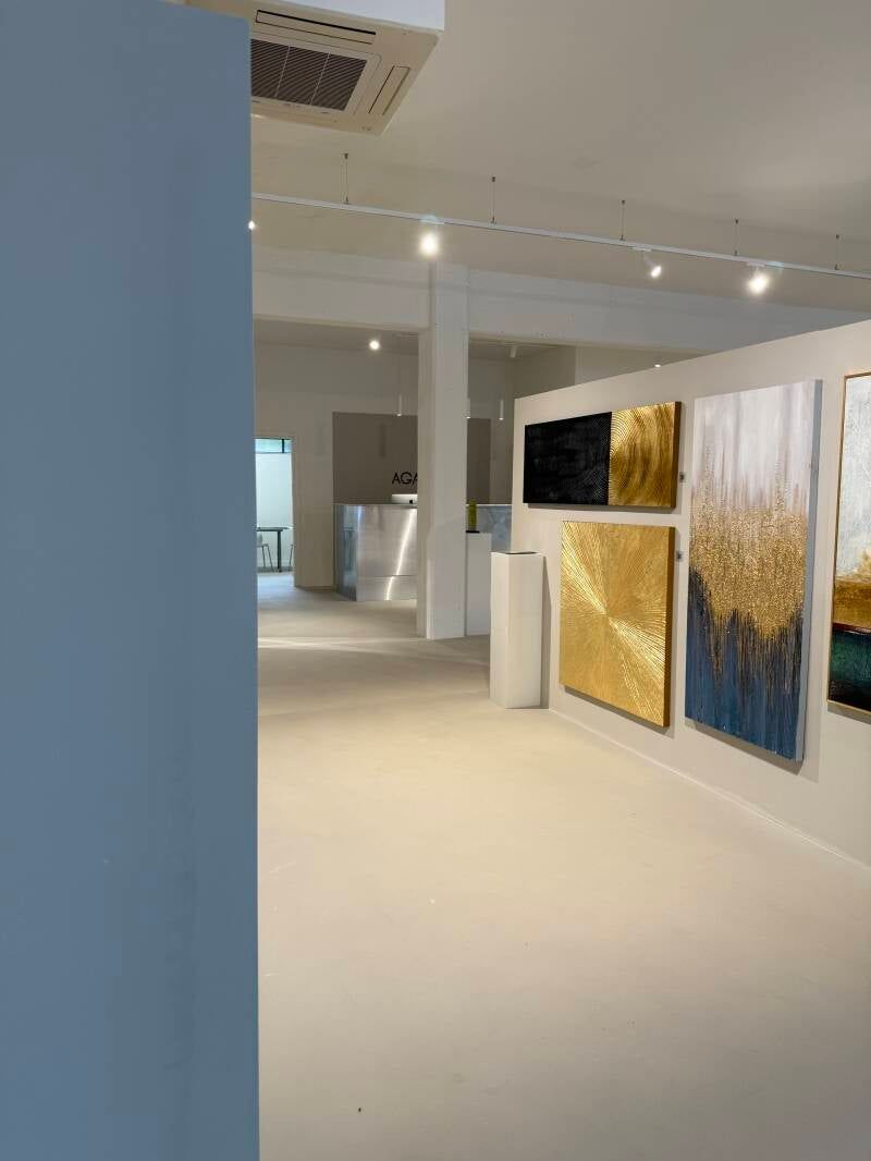 Interno di una galleria d'arte, dipinti esposti, pareti bianche, dettagli dorati e blu, illuminazione intensa.