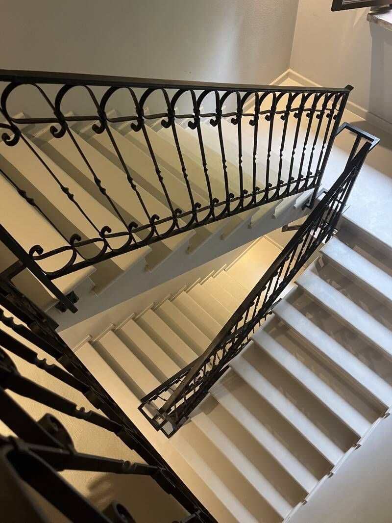 Scala con ringhiera in ferro nero, vista dall'alto. Gradini bianchi, ombre e una parete chiara.