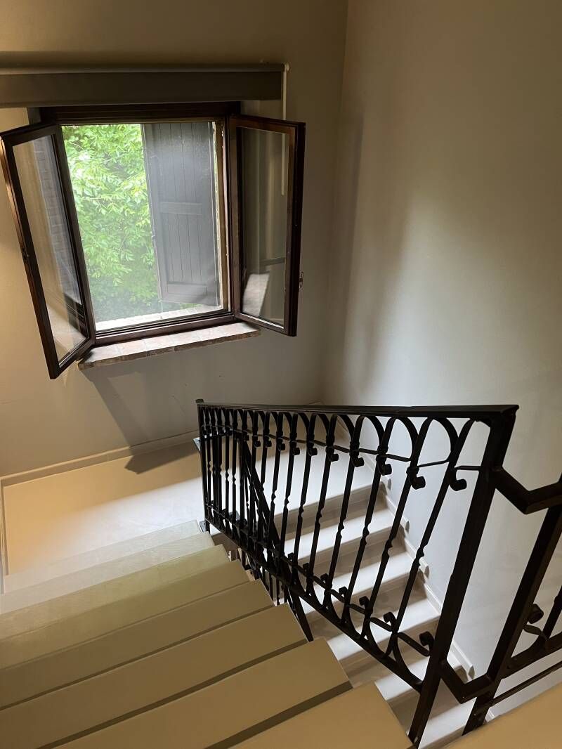 Scala con ringhiera in ferro battuto, gradini bianchi e una finestra aperta che si affaccia sugli alberi.