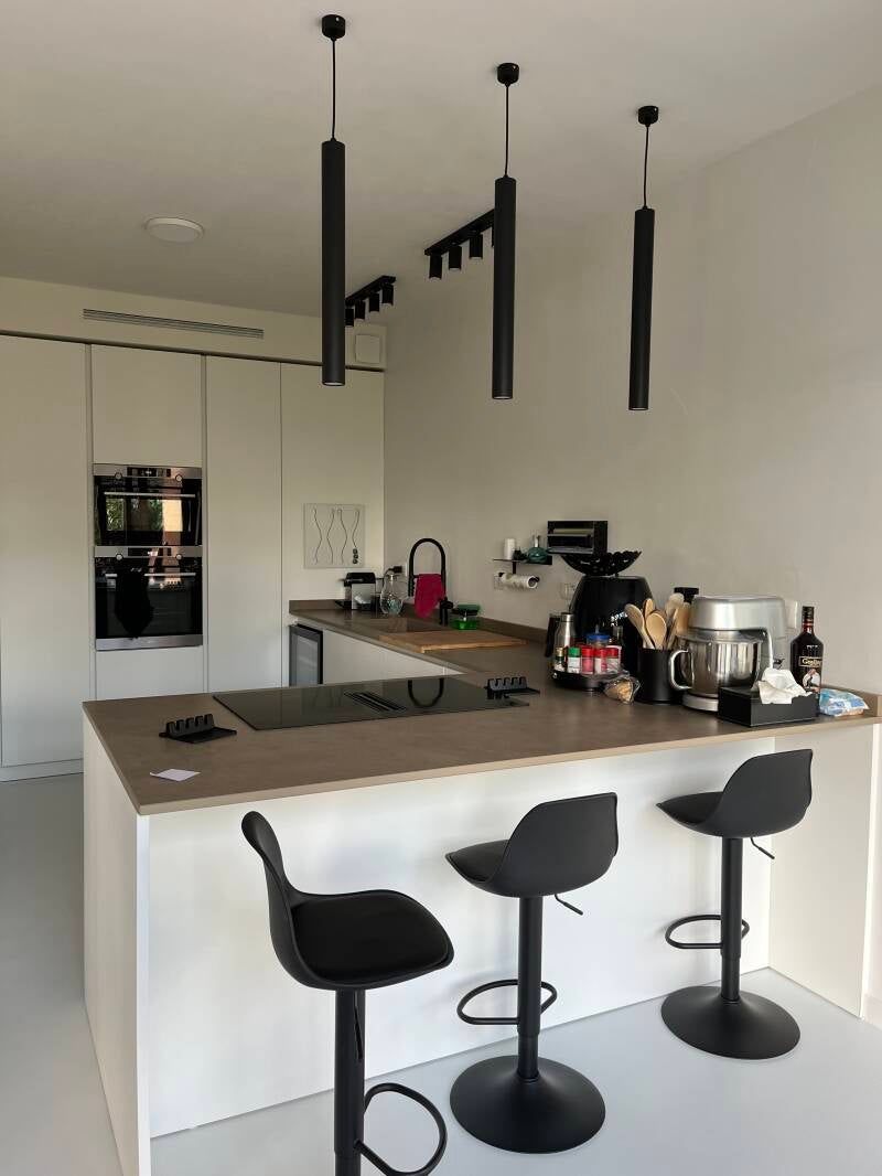 Cucina moderna con bancone per la colazione e tre sgabelli neri. Mobili bianchi, piano di lavoro marrone e lampade a sospensione.