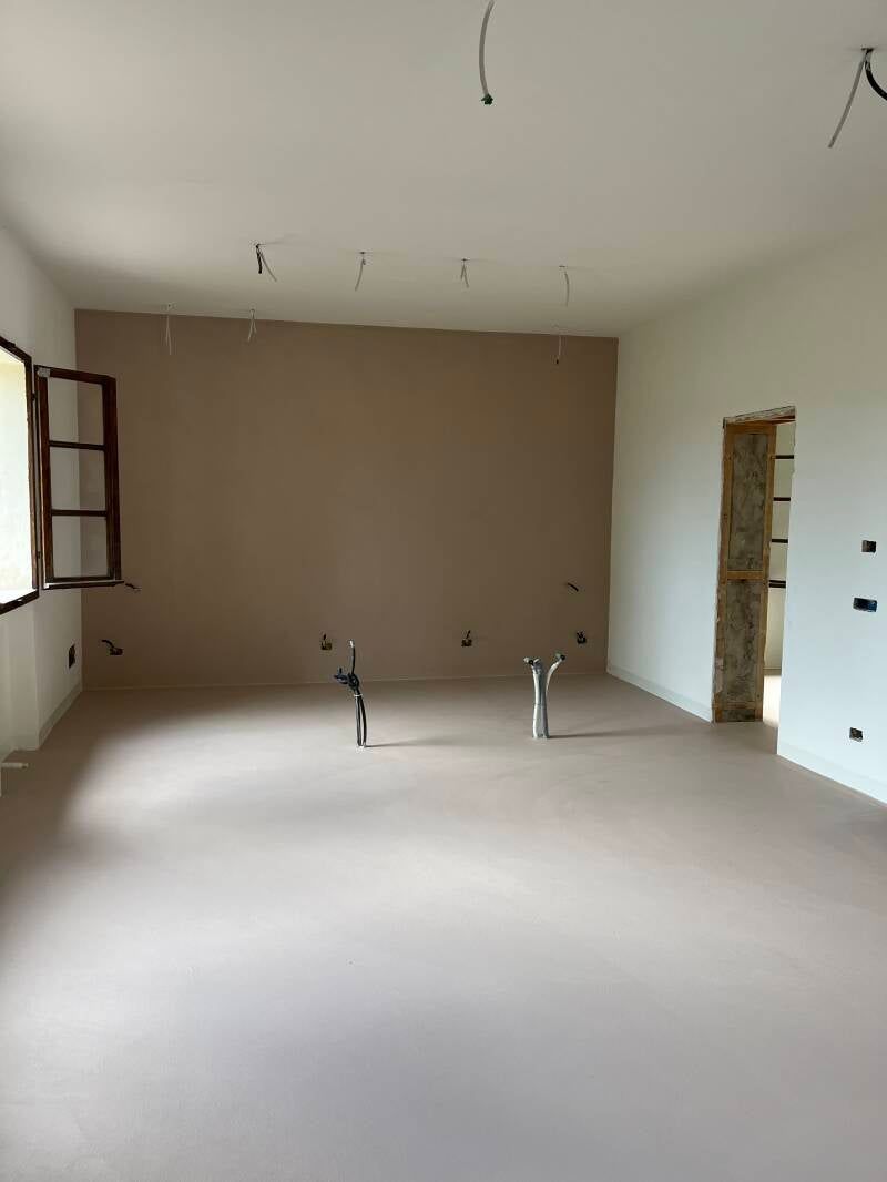 Interno di una stanza in costruzione, con pareti beige e pavimento chiaro. Apparecchi elettrici sporgenti.