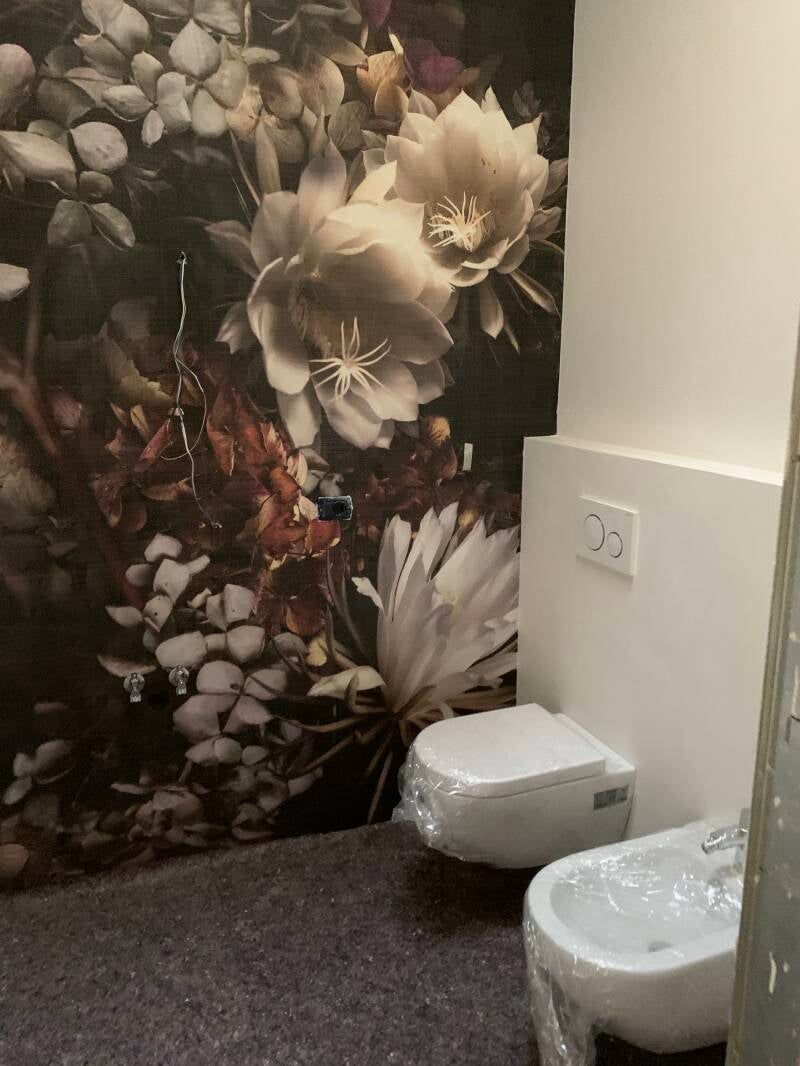 WC e bidet in un bagno con carta da parati floreale e una parete bianca con una presa elettrica.