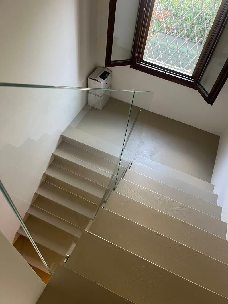 Scala vista dall'alto. Gradini beige scendono da una finestra. Un corrimano è fissato al muro.