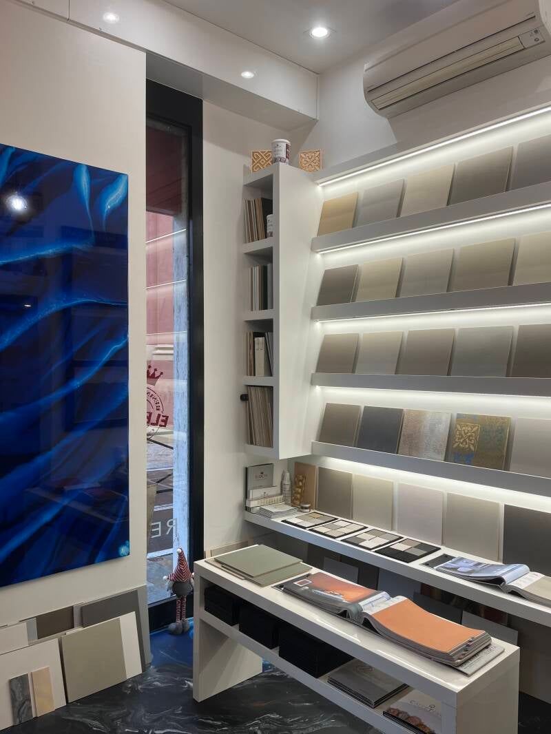 Interno di uno showroom di piastrelle con campioni di piastrelle su scaffali e tavoli. Un'opera d'arte blu è appesa sulla sinistra.