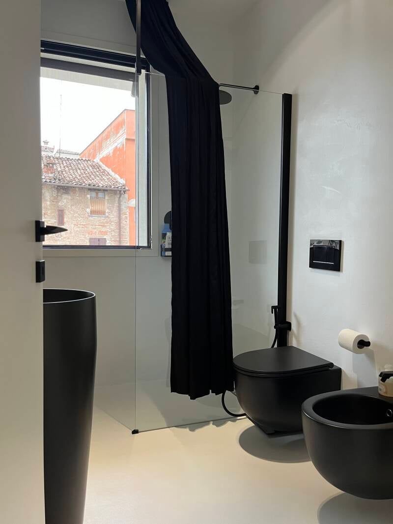 Bagno moderno completamente nero con WC, bidet e lavandino. Tenda da doccia nera davanti alla finestra. Pareti e pavimento bianchi.