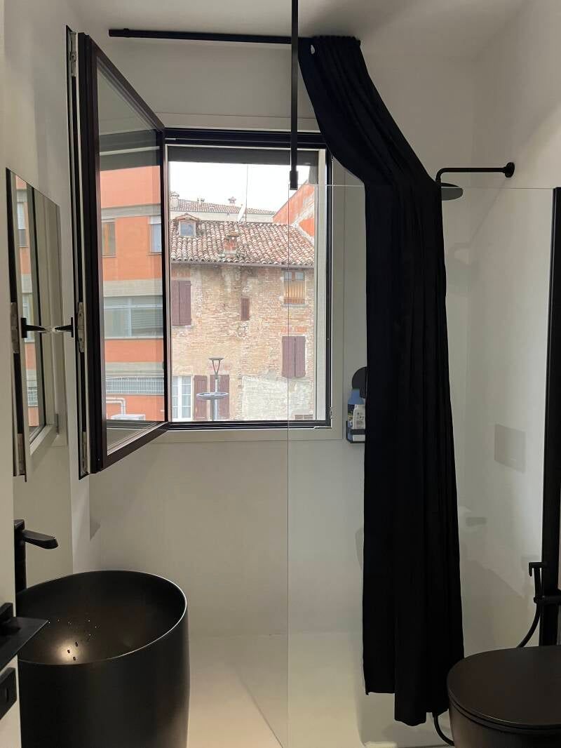 Bagno con sanitari neri, tra cui lavandino, WC e tenda della doccia, accanto a una finestra aperta che si affaccia su un edificio in mattoni.