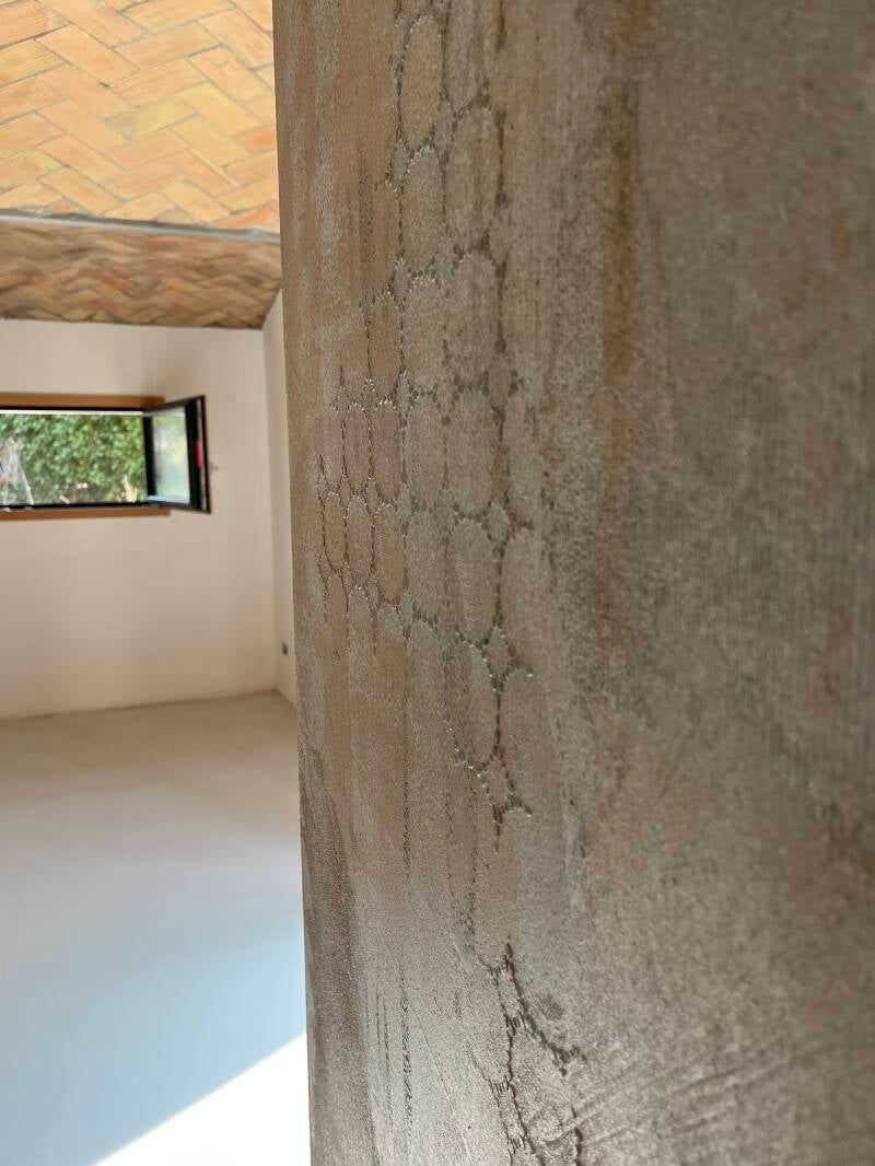 Dettaglio di una parete beige testurizzata in una stanza con pareti bianche, finestra e soffitto in mattoni.