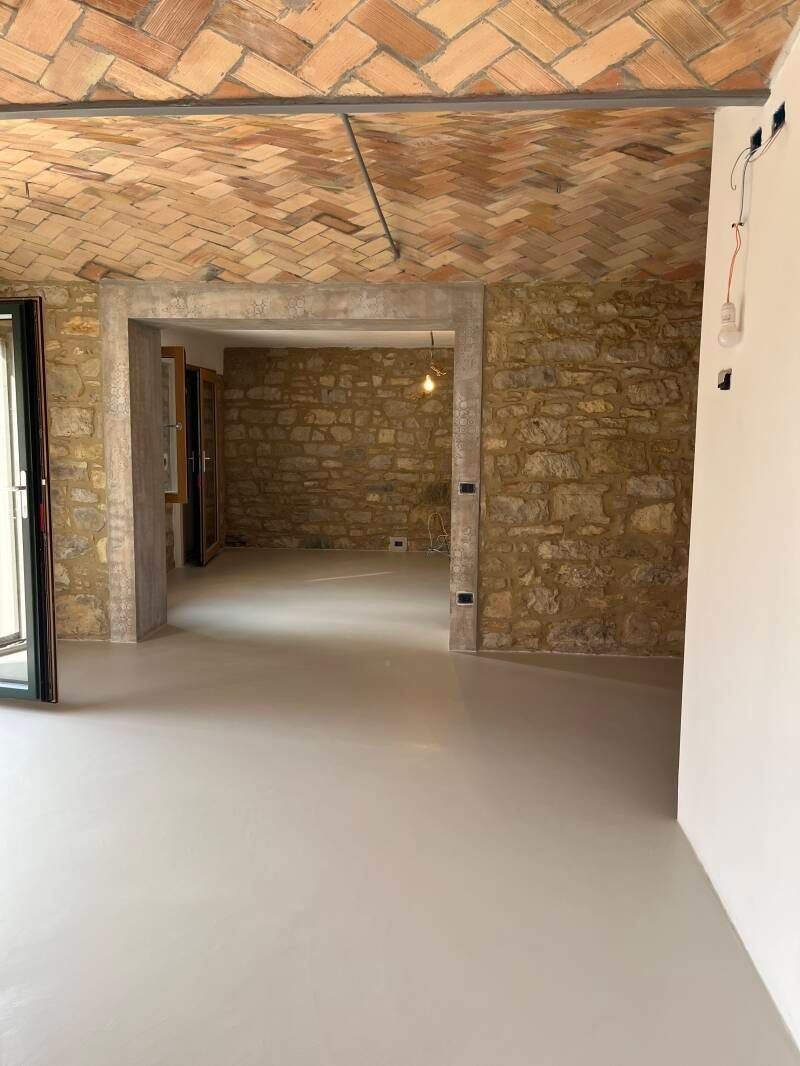 Stanza interna con soffitto in mattoni a vista, pareti in pietra e pavimento grigio chiaro. Porta aperta.