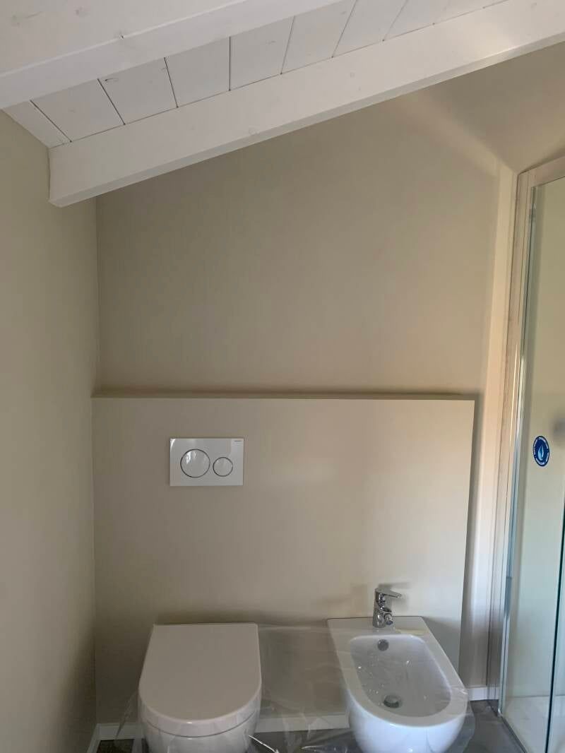 Bagno beige con WC, bidet e doccia. Travi a vista bianche. Placca di comando a muro.