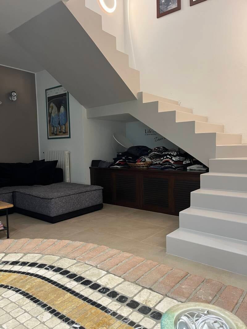 Vista interna con scale, sedute e un tappeto. Dominano i toni del bianco e del marrone. Un mobiletto in legno scuro si trova sotto le scale.