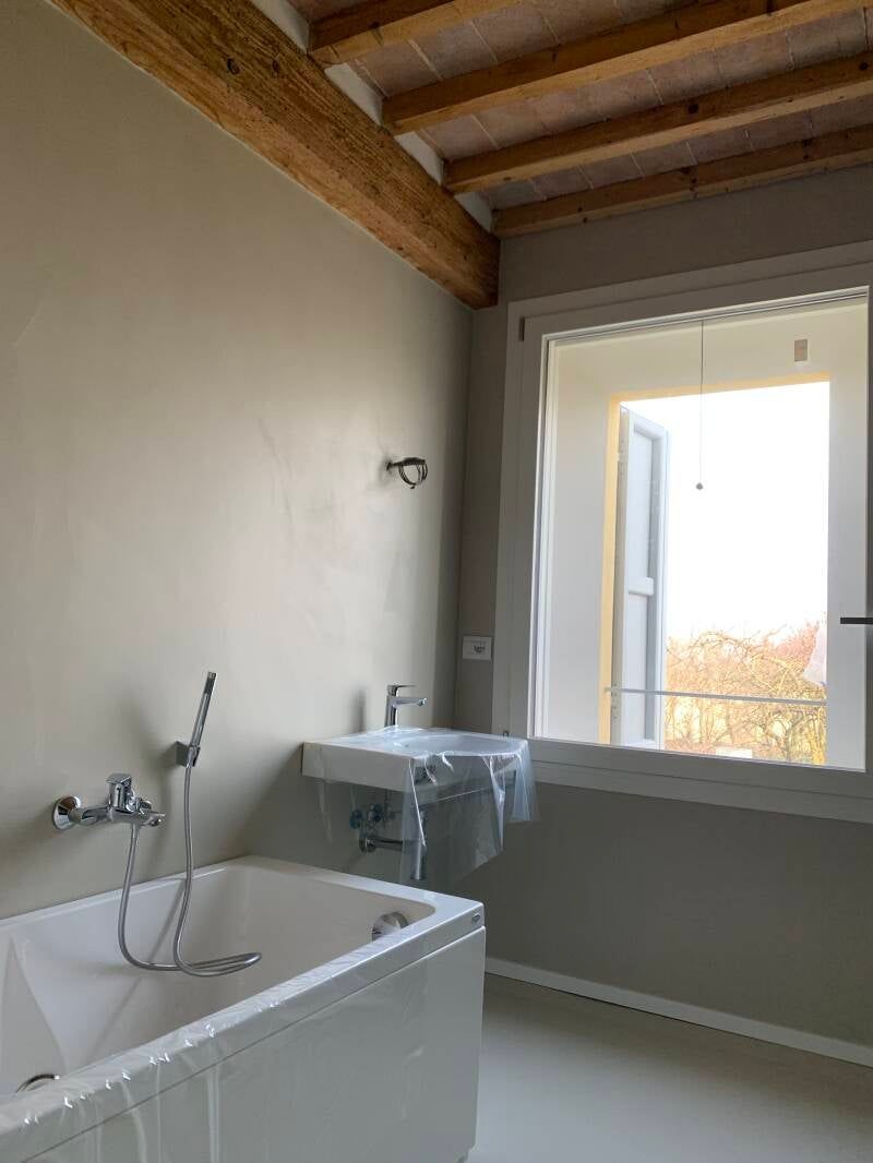 Bagno con vasca bianca, lavandino e finestra; pareti neutre, travi in ​​legno sul soffitto.