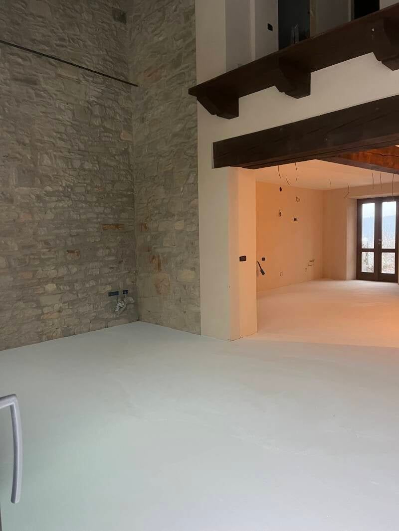 Interno di un edificio con pareti in pietra e bianche, travi a vista e pavimento luminoso; una porta conduce all'esterno.