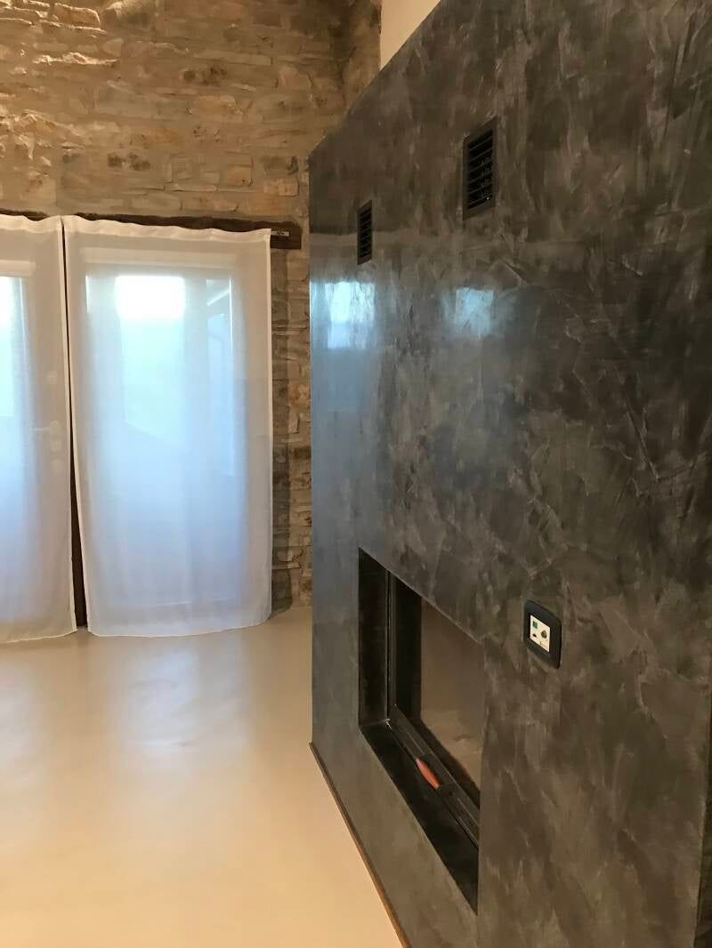 Camino moderno con finitura grigia testurizzata, accanto a tende bianche trasparenti e parete in mattoni.